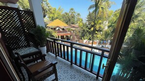 A balcony with a patterned floor, a wooden bench, and a view of a swimming pool and a building with a yellow roof.