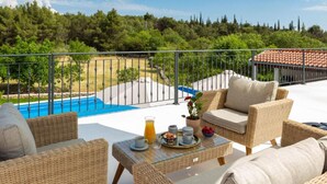 A balcony with wicker furniture, a table with breakfast items, and a pool in the background. - Villa Casa Di Pietra (Sibenik)