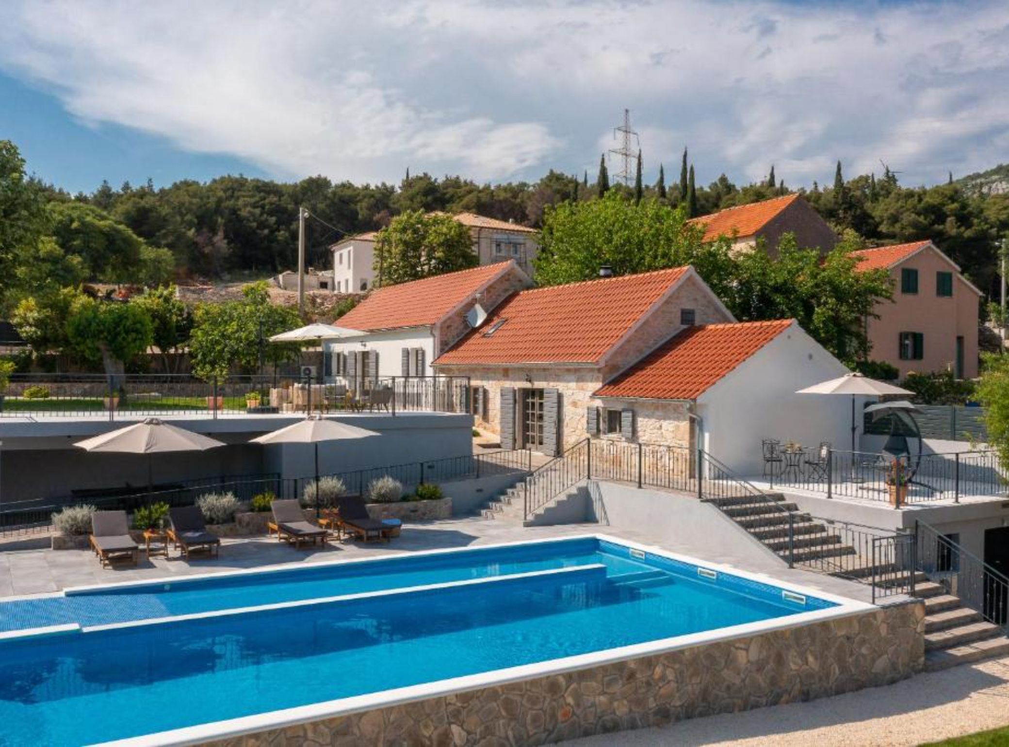 Un espace piscine avec une eau d’un bleu limpide, des marches en pierre menant à un patio couvert, et une vue sur les maisons environnantes, dont les toits sont recouverts de tuiles rouges.
