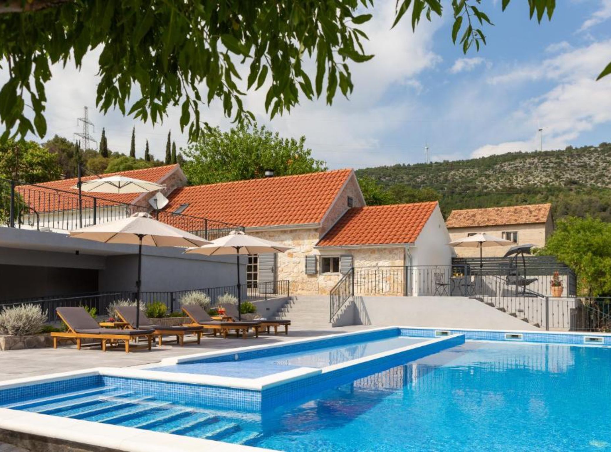 Ein Swimmingpool mit Liegestühlen und Sonnenschirmen, umgeben von Steingeb äuden mit roten Ziegeldächern.