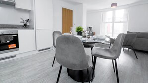 A modern kitchen with white cabinets, a built-in oven, and a dining area with a round table and grey chairs. - Salford - 2 Bed Apartment - Sleeps 4 - Wifi (Salford)