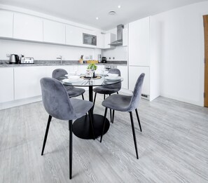 A modern kitchen with white cabinets, a stainless steel range hood, and a dining area with a round table and four grey chairs. - Salford - 2 Bed Apartment - Sleeps 4 - Wifi (Salford)