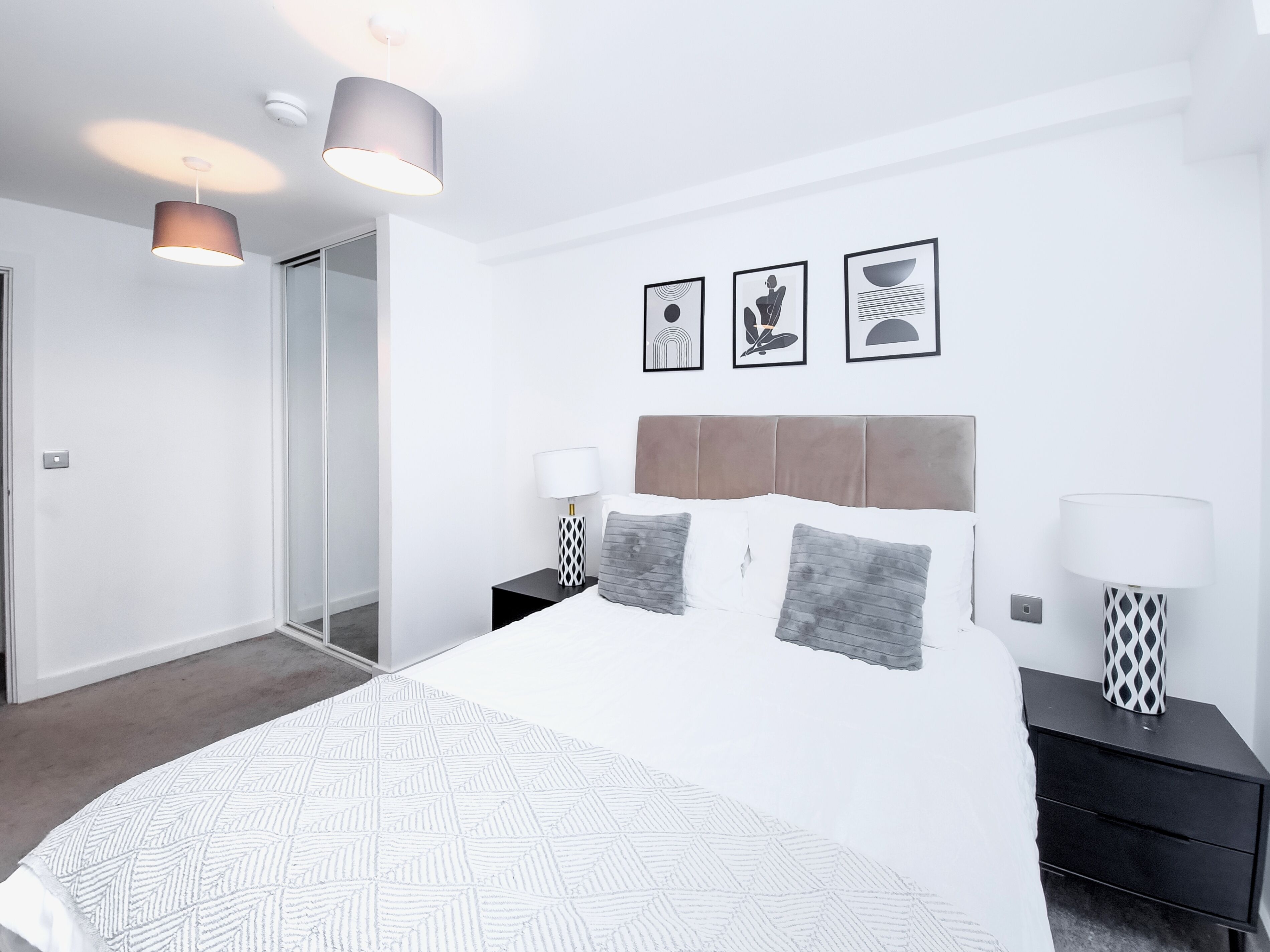 Une chambre moderne avec un grand lit, deux tables de chevet avec des lampes, une tête de lit et trois tableaux encadrés accrochés au mur.