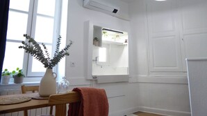 Una habitación luminosa y blanca con ventana, espejo, equipo de aire acondicionado, lámpara colgante y una mesa de madera con un florero y plantas.