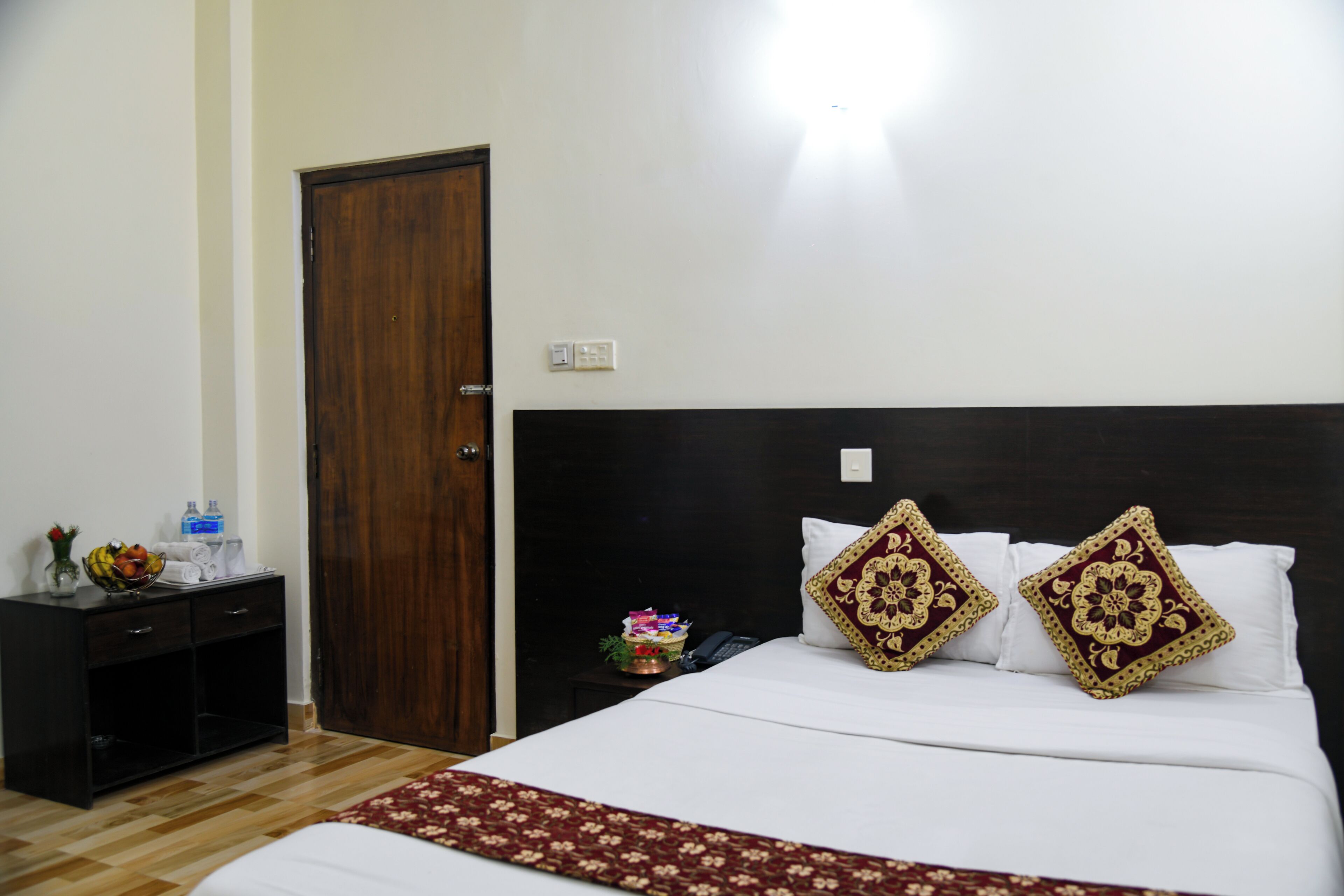 A hotel room with a wooden door, a bedside table with a vase and a bowl of fruit, a bed with white linens and decorative pillows, and a nightstand with a phone and a small plant.