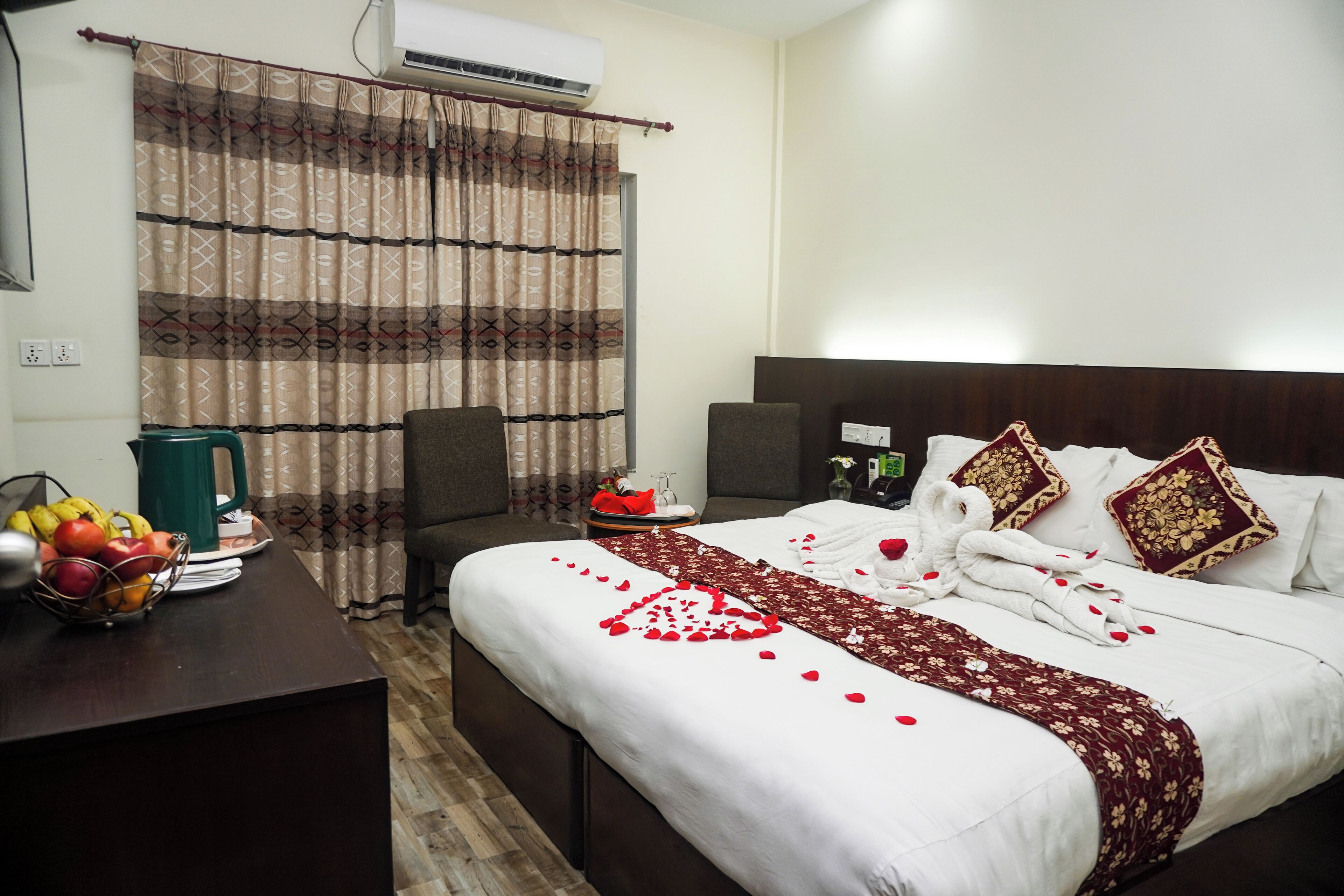 A hotel room with a bed, a desk with fruit, a green kettle, and a curtain.
