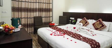 A hotel room with a bed, a desk with fruit, a green kettle, and a curtain.