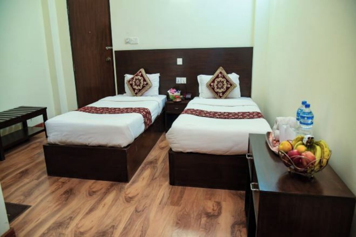 A hotel room with two beds, wooden flooring, a nightstand with a water bottle and fruit, and a small table with a fruit bowl.