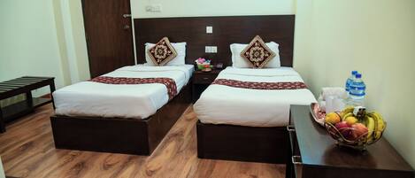 A hotel room with two single beds, wooden flooring, a small table with a fruit basket, and a water bottle on the nightstand.