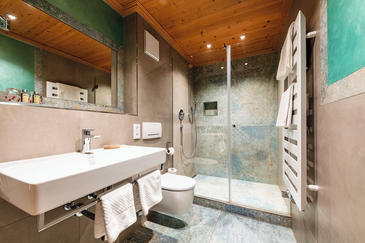 Un bagno moderno con soffitto in legno, uno specchio grande, un lavandino bianco, un water e uno spazio doccia con pareti in vetro.