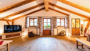 Un amplio ático con vigas de madera a la vista, un ventanal con cortinas florales, un televisor y un mueble de madera.