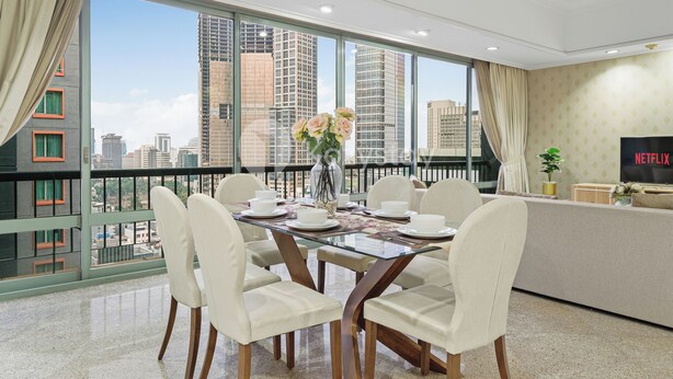 A modern dining area with a glass table, white chairs, a vase of flowers, and a view of tall buildings through large windows. - Brassia by Kozystay - Thamrin (Jakarta)