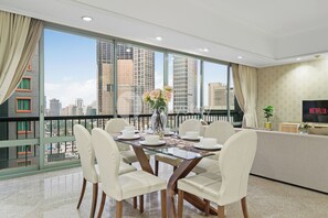 A modern dining area with a glass table, white chairs, a vase of flowers, and a view of tall buildings through large windows. - Brassia by Kozystay - Thamrin (Jakarta)