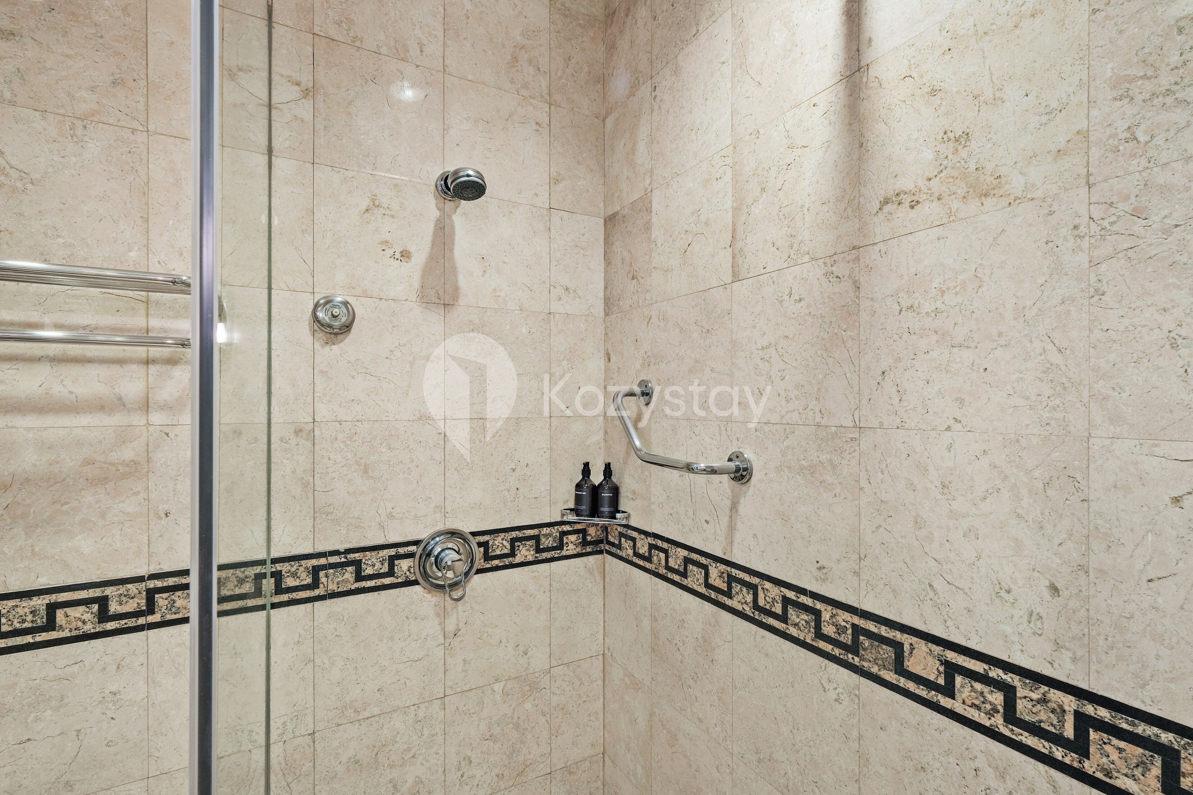 Canto de um banheiro com chuveiro, porta de vidro e borda de azulejos decorativos.