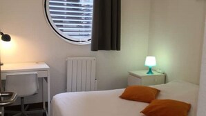 Una camera d'albergo con un letto, una scrivania, una sedia, una lampada e una finestra con persiane.