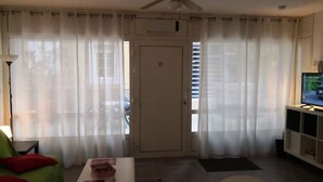 Un soggiorno con ventilatore a soffitto, televisore su un mobile, una porta con un cartello e tende leggere.