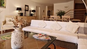 Una sala de estar moderna con un sofá blanco, una mesa de centro de vidrio y una silla de mimbre. Hay un jarrón grande con flores en el suelo, y se ve una escalera al fondo.