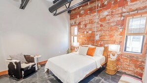 Ein Schlafzimmer mit Backsteinwand, einem Bett mit weißer Bettwäsche, einem kleinen Nachttisch und einer Couch mit einem gemusterten Kissen.