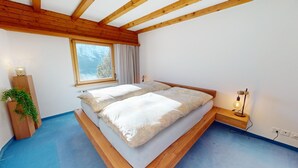 Una camera da letto con soffitto in legno, un'ampia finestra, un letto con lenzuola bianche, un comodino con una lampada e una piccola pianta.