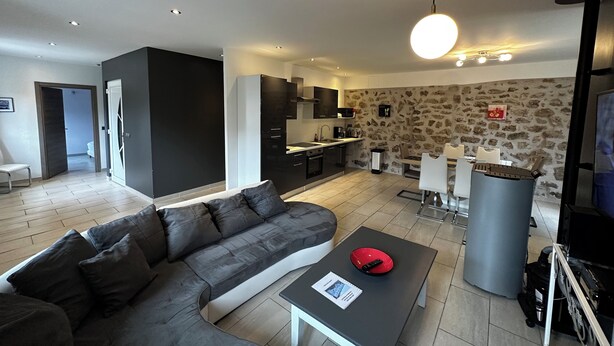 Ein modernes Wohnzimmer mit einem grauen Ecksofa, einem kleinen Tisch mit Fernbedienung und einer Küchenzeile mit Wandverkleidung aus Stein.