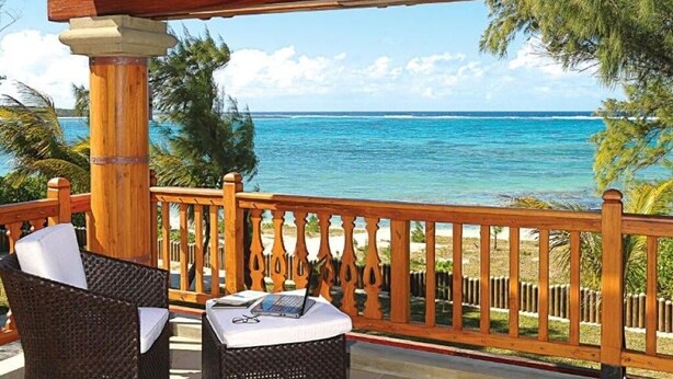 Un balcone in legno con mobili in vimini e un portatile, con vista su una spiaggia con acqua cristallina e palme.