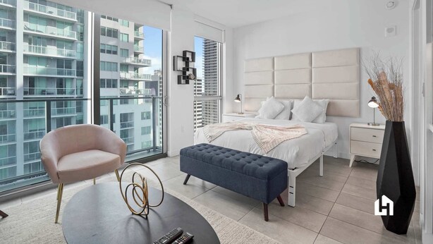 Un dormitorio moderno con una cama grande, mesitas de noche, una silla y vistas a la ciudad a través de ventanales.
