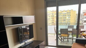 Un salón moderno con un televisor de pantalla plana, un mueble de entretenimiento oscuro, un sofá marrón y un ventanal corredizo que da a un balcón exterior con vistas a edificios amarillos.