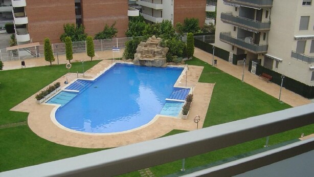 Una piscina rectangular con una pequeña cascada, rodeada de un césped cuidado y algunos árboles.