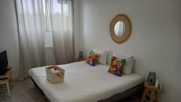 Une chambre d’hôtel bien rangée, avec un grand lit, une table de chevet avec une lampe, un petit tabouret et un téléviseur posé sur un bureau.