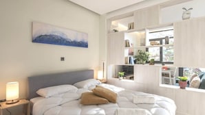 A modern bedroom with a large bed, a bedside table with a lamp, a painting on the wall, and a built-in shelving unit with books and plants. - Luxury Haven in the most ideal area of Athens (Athina)