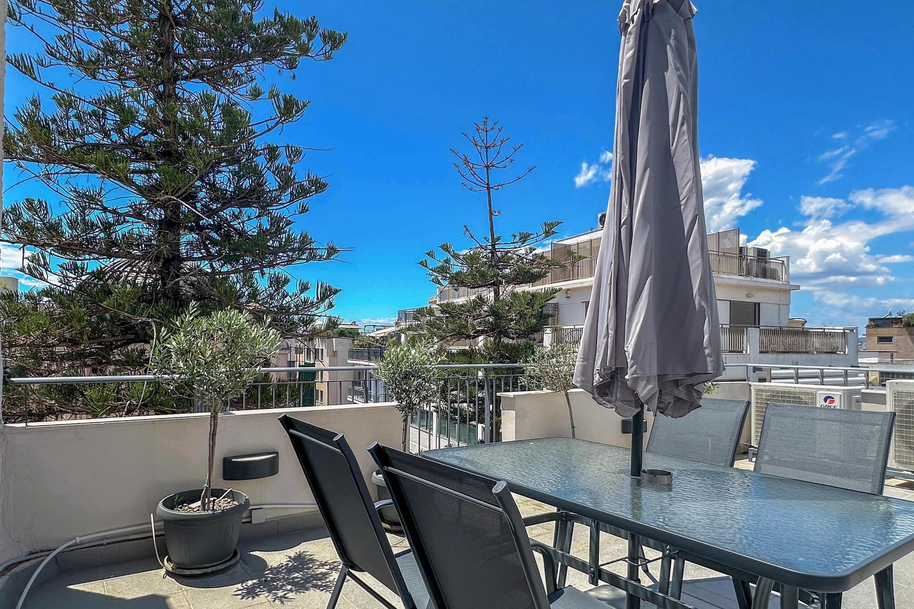 A rooftop terrace with a table and chairs, a potted tree, and a grey umbrella.