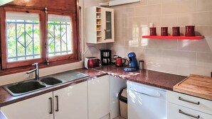 A kitchen with white cabinets, a sink, a window with a view of greenery, a coffee maker, and a red shelf with cups. - Sueño II by Interhome (L'Ametlla de Mar)
