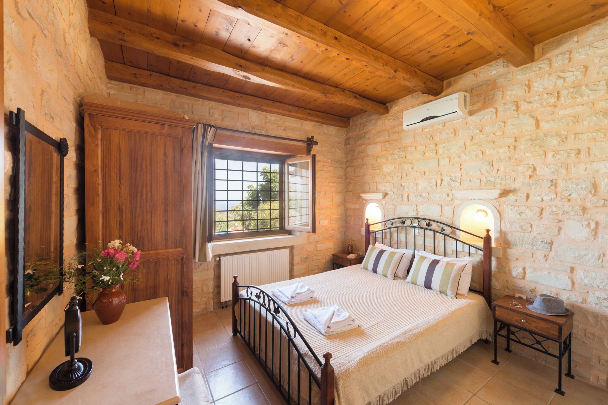 Een slaapkamer met een stenen muur, houten plafond, een bed met kussens, een nachtkastje, een vaas met bloemen en een raam met uitzicht.