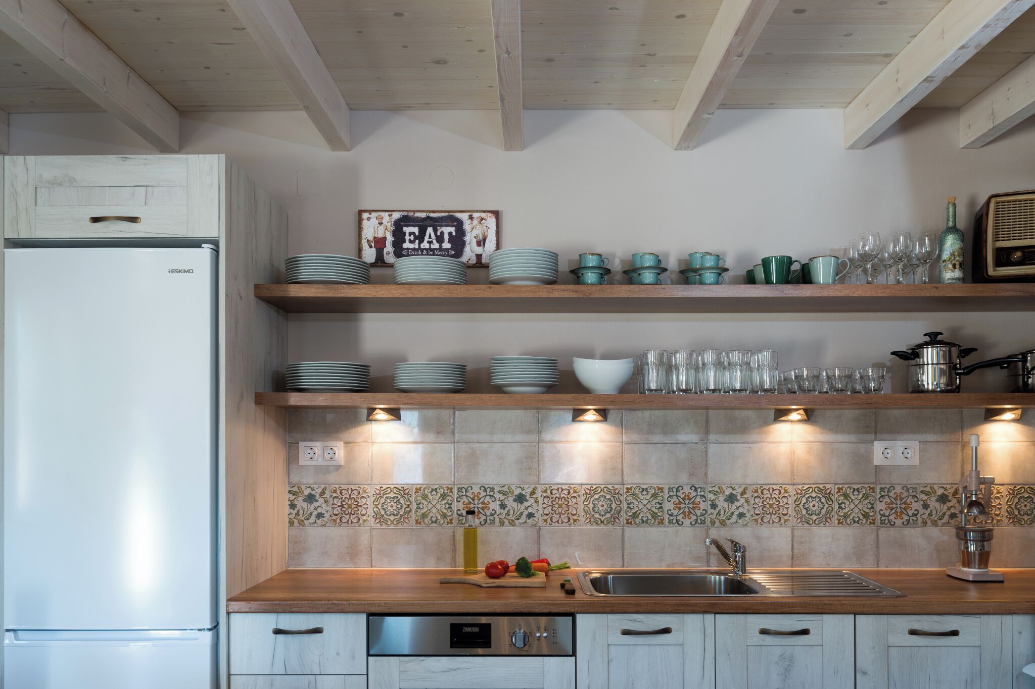 Una cocina con armarios blancos, un refrigerador de acero inoxidable, estantes de madera con vajilla y un salpicadero de azulejos.