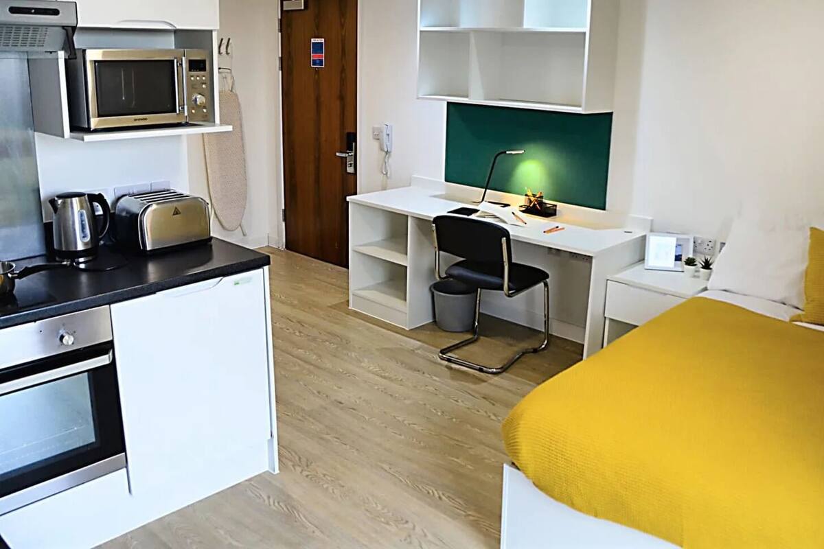 A compact hotel room with a kitchenette, a bed with a yellow bedspread, a desk with a chair, and a television.