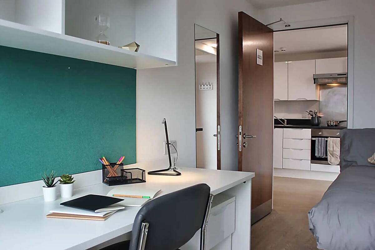 A modern hotel room with a white desk, a green accent wall, a black chair, a bed, and a kitchenette in the background.