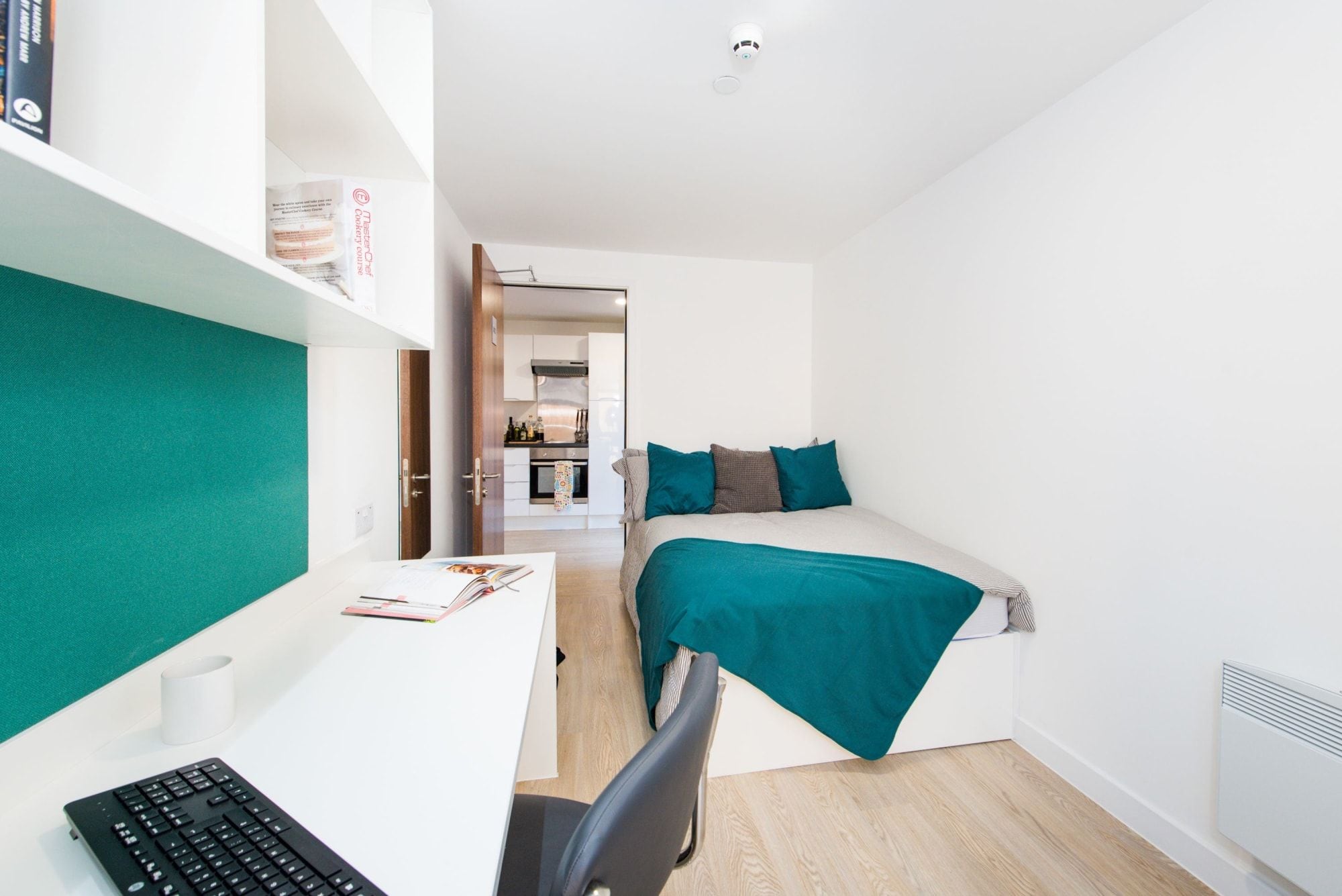 A compact room with a bed, a desk with a keyboard, and a chair. There is a kitchenette in the background.