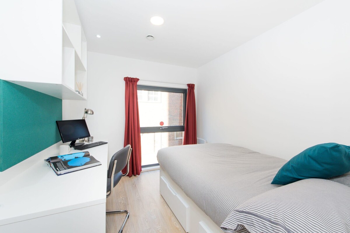 A bedroom with a bed, a desk with a computer, a chair, and a window with curtains.
