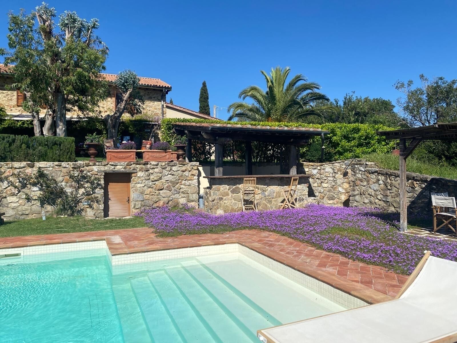 A pool area with a stone wall, a pergola, and purple flowers.