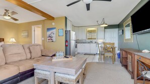 A living room with a sofa, a wooden table, and a dining area with chairs. There is a kitchen with white cabinets and a refrigerator. - Beach & Tennis 1802 (Bonita Springs)