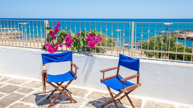 Dos sillones de director de madera con cojines azules en un balcón con vista al mar.