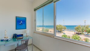Una habitación con un gran ventanal con vistas a la playa, un escritorio con una computadora portátil, una silla y un cuadro con un velero.