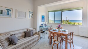Una sala de estar moderna con sofá, mesa de comedor y sillas, vista al mar a través de ventanas amplias y obras de arte en las paredes.
