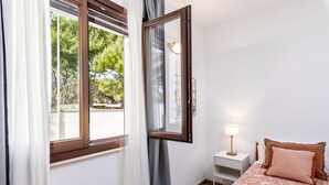 Un dormitorio con cama, mesita de noche, lámpara, ventana con cortinas y vista a árboles.