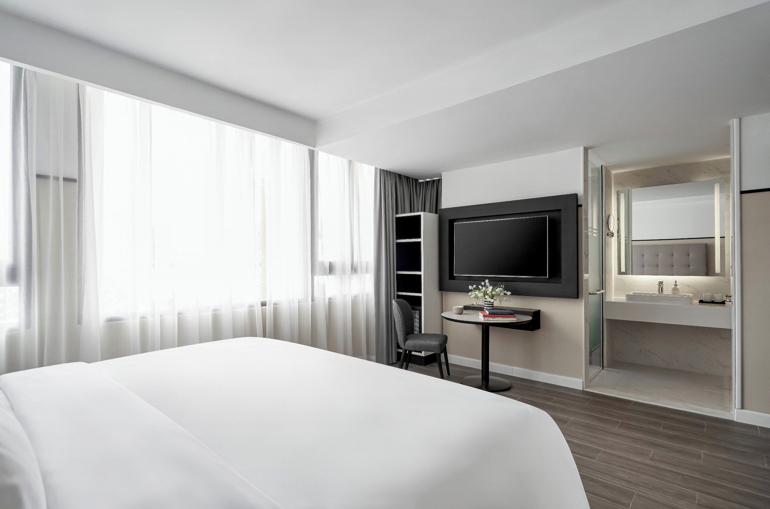 Una habitación de hotel moderna con una cama grande, un escritorio con una silla, un televisor montado en la pared y un baño con tocador y espejo.
