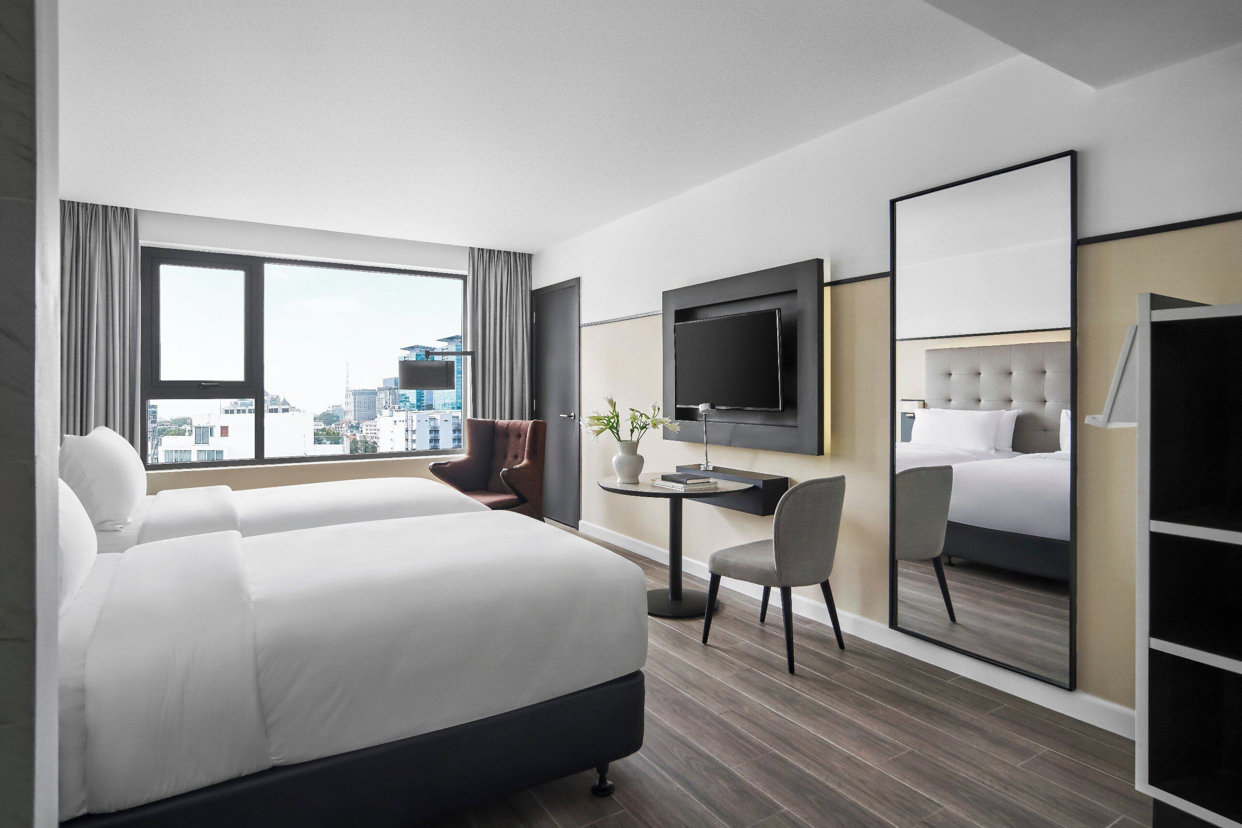 A modern hotel room with a large bed, a desk, a chair, a TV, and a large window with a city view.