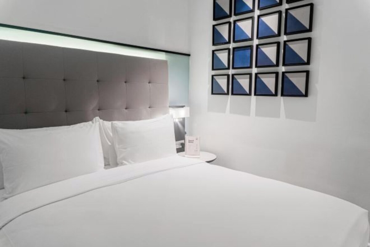 Une chambre à coucher moderne avec une tête de lit capitonnée, un lit blanc, une table de chevet avec une lampe et un mur orné d’œuvres d’art géométriques encadrées.