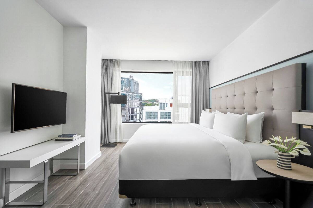 Une chambre d’hôtel moderne dotée d’un grand lit, d’un téléviseur à écran plat, d’une table de chevet et offrant une vue sur la ville grâce à de grandes fenêtres.