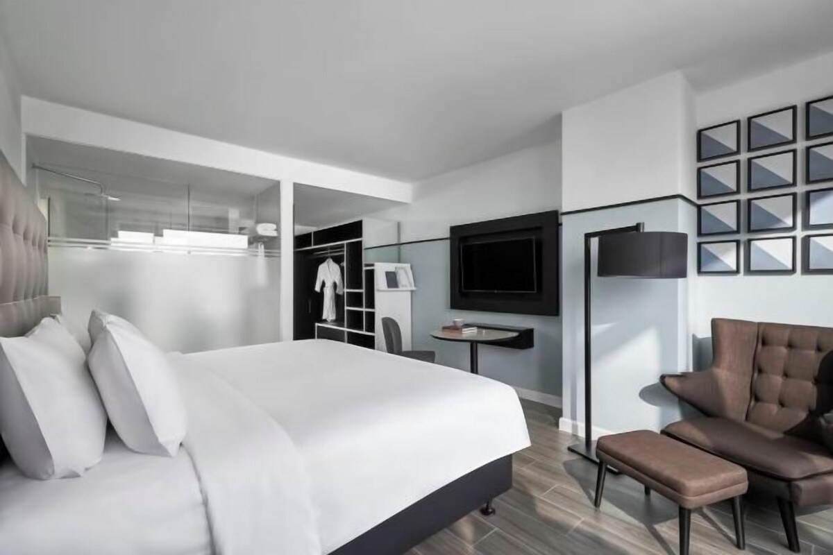 A modern hotel room with a large bed, a seating area with a sofa and armchair, a TV, a wardrobe, and a mirror.