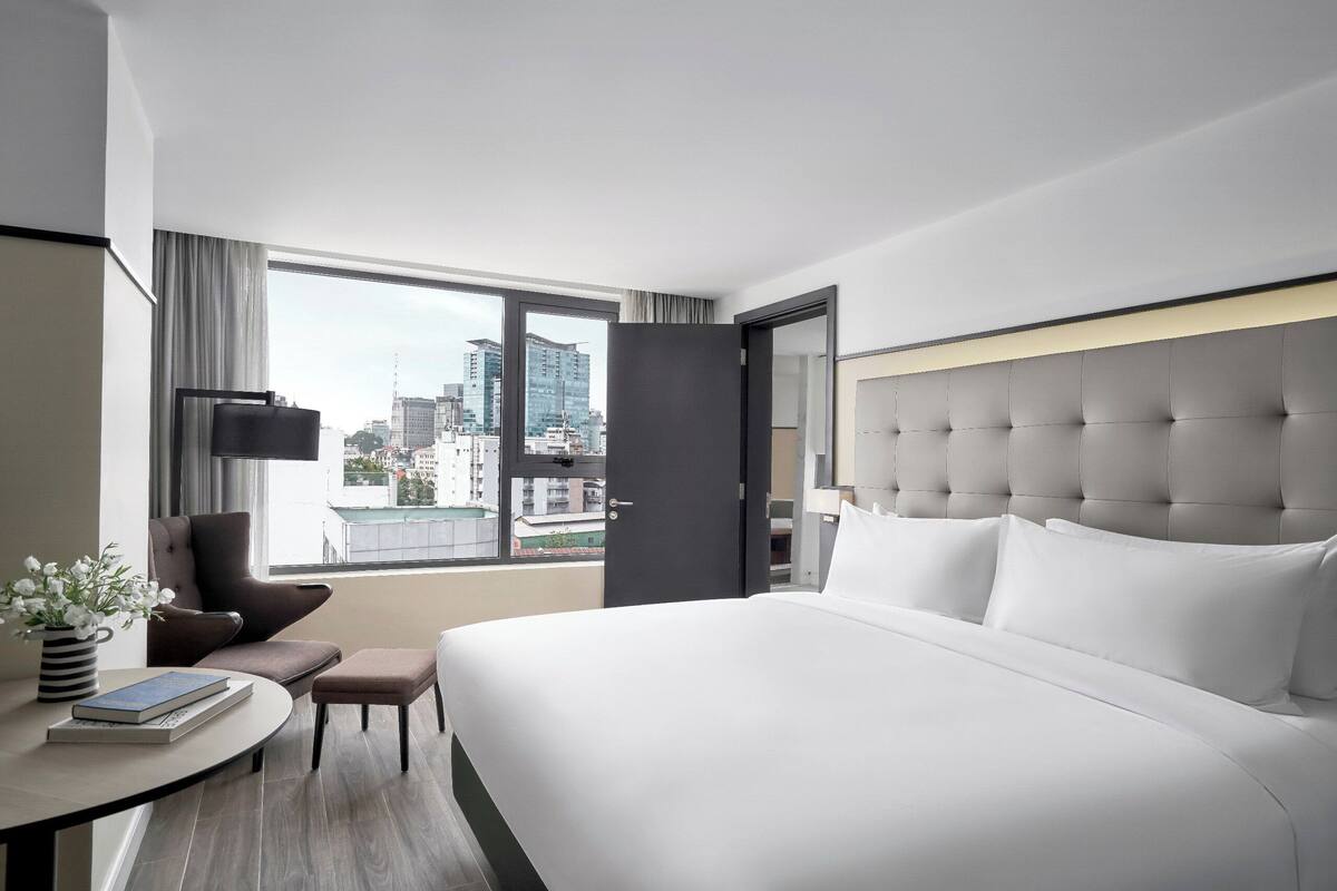 Une chambre d’hôtel moderne avec un grand lit, une petite table sur laquelle sont posés un livre et un vase de fleurs, une chaise, et une vue sur la ville par une grande fenêtre.